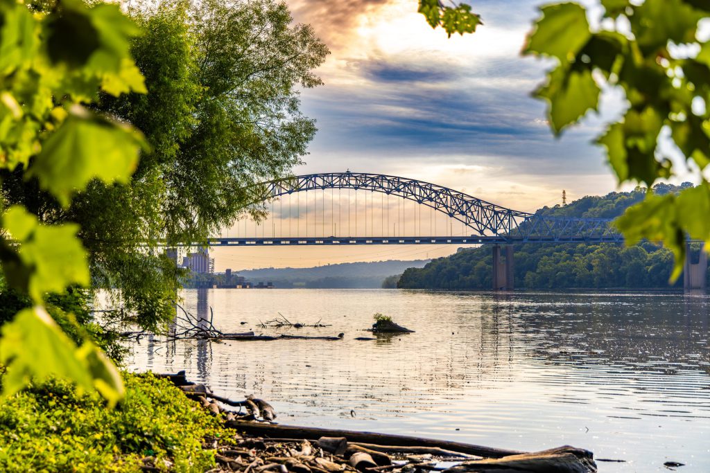 A steel arch bridge over the Ohio River at sunset, framed by leaves