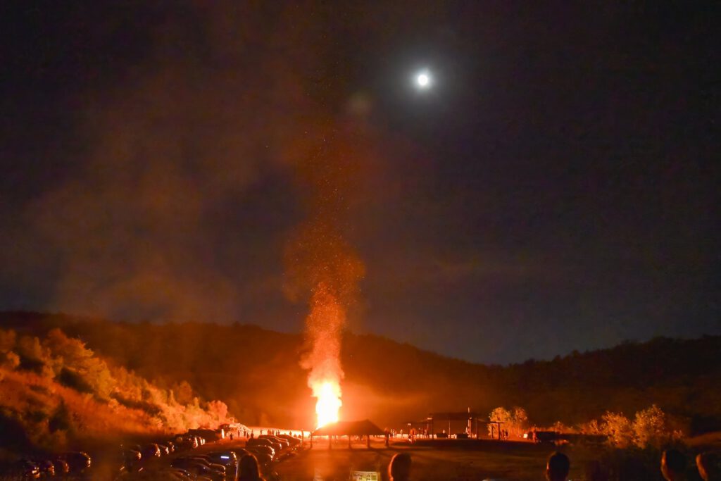 A tall bonfire throws sparks into the night sky beside a pavilion, with the moon above the hills